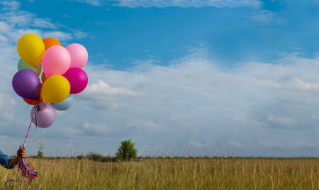 Woman holding balloons running on green meadow white cloud and blue sky with happiness Cheerful and relax. Hands holding vibrant air balloons play on birthday party happy times summer sunlight outdoorの写真素材