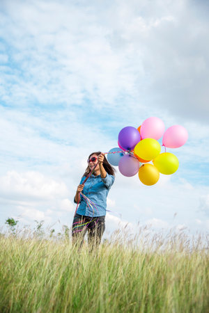 Woman holding balloons running on green meadow white cloud and blue sky with happiness Cheerful and relax. Hands holding vibrant air balloons play on birthday party happy times summer sunlight outdoorの写真素材