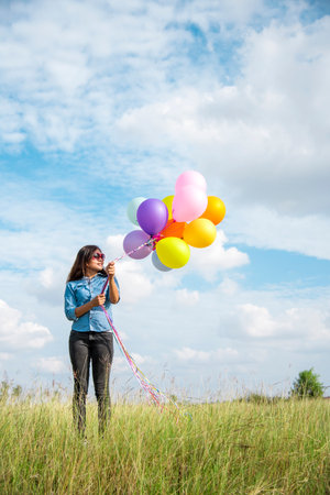 Woman holding balloons running on green meadow white cloud and blue sky with happiness Cheerful and relax. Hands holding vibrant air balloons play on birthday party happy times summer sunlight outdoorの写真素材