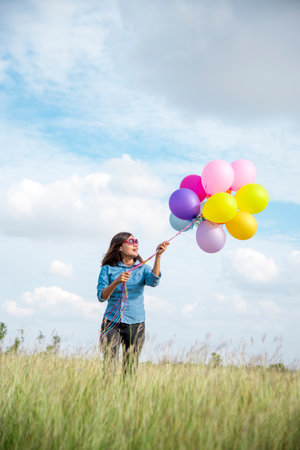 Woman holding balloons running on green meadow white cloud and blue sky with happiness Cheerful and relax. Hands holding vibrant air balloons play on birthday party happy times summer sunlight outdoorの写真素材