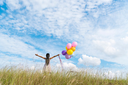 Cheerful cute girl holding balloons running on green meadow white cloud and blue sky with happiness. Hands holding vibrant air balloons play on birthday party happy times summer on sunlight outdoorの写真素材