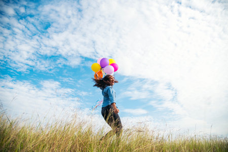 Cheerful cute girl holding balloons running on green meadow white cloud and blue sky with happiness. Hands holding vibrant air balloons play on birthday party happy times summer on sunlight outdoorの写真素材