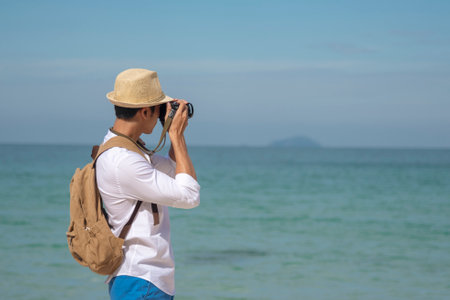 Man traveler using digital camera on summer beach blue sky take a photo. Asian Photographer man journey relax on the beach in summertime. Man backpack shooting photo asia destination holiday tripの写真素材