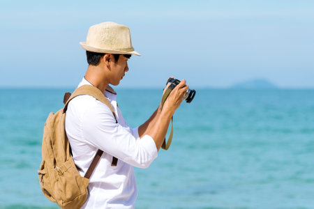Man traveler using digital camera on summer beach blue sky take a photo. Asian Photographer man journey relax on the beach in summertime. Man backpack shooting photo asia destination holiday tripの写真素材