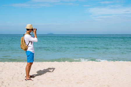 Man traveler using digital camera on summer beach blue sky take a photo. Asian Photographer man journey relax on the beach in summertime. Man backpack shooting photo asia destination holiday tripの写真素材