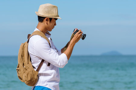 Man traveler using digital camera on summer beach blue sky take a photo. Asian Photographer man journey relax on the beach in summertime. Man backpack shooting photo asia destination holiday tripの写真素材