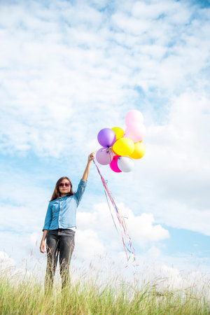 Woman holding balloons running on green meadow white cloud and blue sky with happiness Cheerful and relax. Hands holding vibrant air balloons play on birthday party happy times summer sunlight outdoorの写真素材