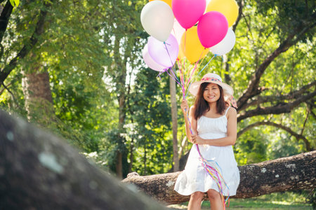 Cheerful beauty woman holding balloons relax sitting under big tree in green park with happiness. Woman Hands holding vibrant air balloons play on birthday party happy time summer on sunshine outdoorの写真素材