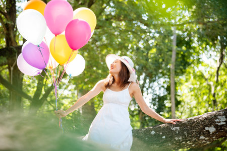 Cheerful beauty woman holding balloons relax sitting under big tree in green park with happiness. Woman Hands holding vibrant air balloons play on birthday party happy time summer on sunshine outdoorの写真素材