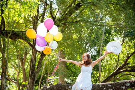 Cheerful beauty woman holding balloons relax sitting under big tree in green park with happiness. Woman Hands holding vibrant air balloons play on birthday party happy time summer on sunshine outdoorの写真素材
