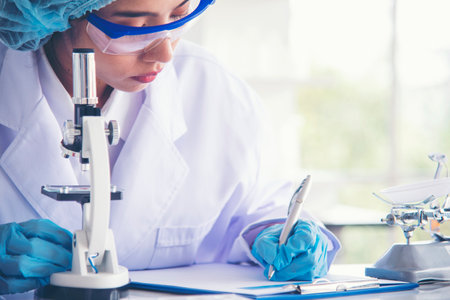 Asian Woman scientist hand write note research in medical lab look at science microscope medical test. Researcher biology chemistry. Asian Female technician laboratory using microscope blood testの写真素材