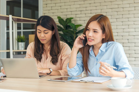 Two businesswomen team meeting using laptop at company office desk. Two young Female freelance reading financial graph charts Planning analyzing marketing data. Asian people team working office firm.の写真素材