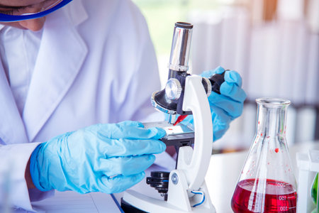 Woman scientist hand write note information in medical lab look at science microscope medical test, research biology chemistry. Asian Females technician laboratory analyzing blood test pharmaceuticalの写真素材