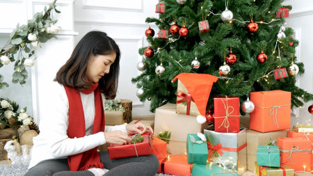 Woman wrapping christmas gift box under xmas tree. Asian woman decor bauble ball christmas tree in xmas holiday winter seasonal celebration. Close up woman hands wrap present gift in cozy living roomの写真素材