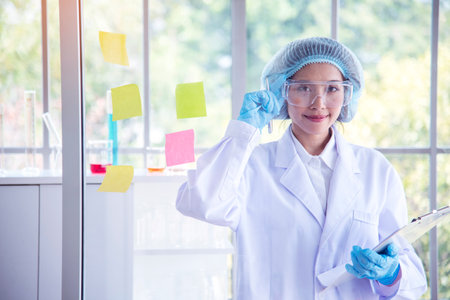 Woman scientist hand write note information in medical lab look at science microscope medical test, research biology chemistry. Asian Females technician laboratory analyzing blood test pharmaceuticalの写真素材