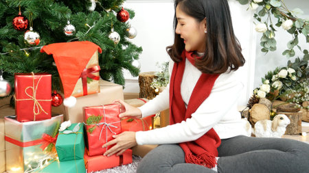 Woman wrapping christmas gift box under xmas tree. Asian woman decor bauble ball christmas tree in xmas holiday winter seasonal celebration. Close up woman hands wrap present gift in cozy living roomの写真素材