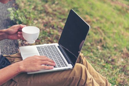 Man hands typing laptop notebook holding coffee cup outside office. Close up man hands using computer laptop sit on green park outdoors lifestyle. freelancer Men using laptop home office technologyの写真素材
