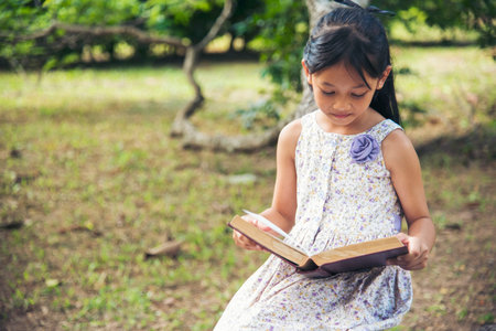 Asian Girl holding book reading at green park in natural garden. Young todler girl relaxation read open book self study. Happy child women smiling with happiness learning. Kid sit in green parkの写真素材