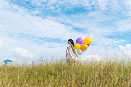 Cheerful cute girl holding balloons running on green meadow white cloud and blue sky with happiness. Hands holding vibrant air balloons play on birthday party happy times summer on sunlight outdoorの写真素材