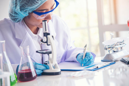 Asian Woman scientist hand write note research in medical lab look at science microscope medical test. Researcher biology chemistry. Asian Female technician laboratory using microscope blood testの写真素材