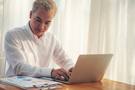 Asian man freelance using laptop at home office desk. man reading financial graph chart Planning analyzing computer data. Asian male people working office business stuff, coffee cup at home officeの写真素材