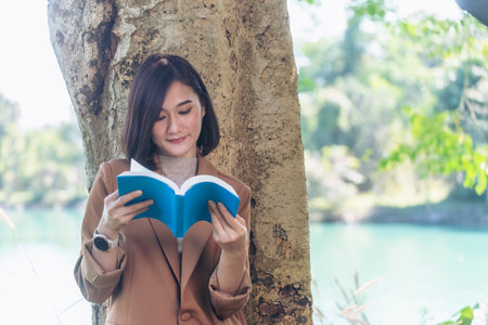 Happy asian woman reading book outdoor in green park nature. Reading book magazine relaxation woman. Leisure portrait woman smile sitting in green park read e-book diary planner with happy faceの写真素材