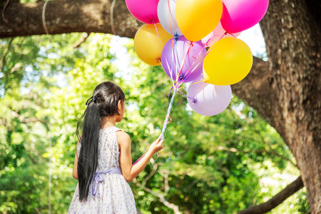 Cheerful cute girl holding balloons running on green meadow white cloud and blue sky with happiness. Hands holding vibrant air balloons play on birthday party happy times summer on sunlight outdoorの写真素材
