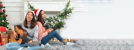 Banner happy asian woman sitting Christmas gift box xmas tree play acoustic guitar together. Banner two women smile sitting christmas ball xmas holiday winter festival in living room with copy spaceの写真素材