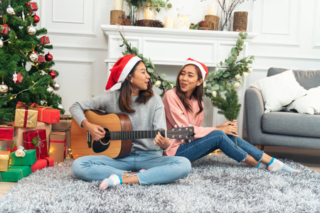 Happy Asian Woman sitting Christmas gift box under xmas tree playing acoustic guitar together. Two Women smile talk sitting decor christmas ball tree xmas holiday winter festival in living roomの写真素材