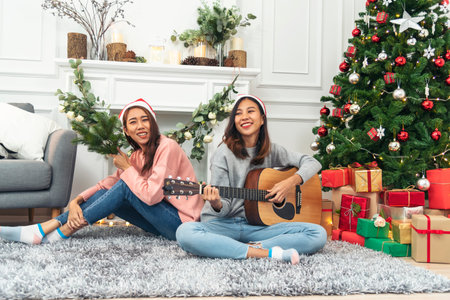 Happy Asian Woman sitting Christmas gift box under xmas tree playing acoustic guitar together. Two Women smile talk sitting decor christmas ball tree xmas holiday winter festival in living roomの写真素材