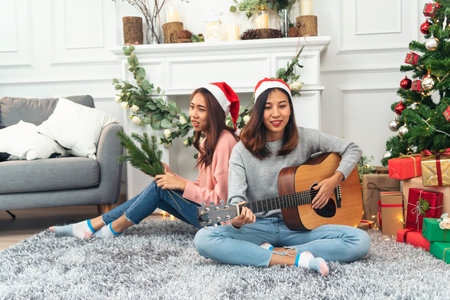 Happy Asian Woman sitting Christmas gift box under xmas tree playing acoustic guitar together. Two Women smile talk sitting decor christmas ball tree xmas holiday winter festival in living roomの写真素材