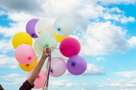 Cheerful cute girl holding balloons running on green meadow white cloud and blue sky with happiness. Hands holding vibrant air balloons play on birthday party happy times summer on sunlight outdoorの写真素材