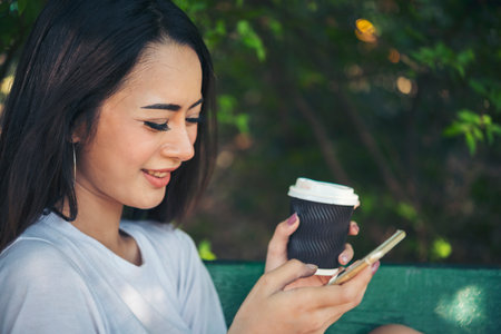 Asian Woman use smartphone drinking coffee hand holding hot disposable cup in green park. Banner Happy Relax asian woman smiling face at outdoors garden. Young women enjoy nature with copy spaceの写真素材