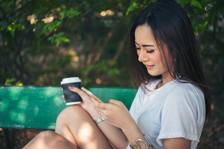 Asian Woman use smartphone drinking coffee hand holding hot disposable cup in green park. Banner Happy Relax asian woman smiling face at outdoors garden. Young women enjoy nature with copy spaceの写真素材