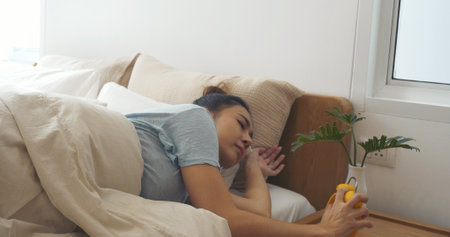 Asian women sleep wake up in white cozy bedroom happy time morning sunrise. Beautiful chinese use hand stop time alarm clock. Happy person lifestyle wake up in morning wellbeing relax sleepy nappingの写真素材