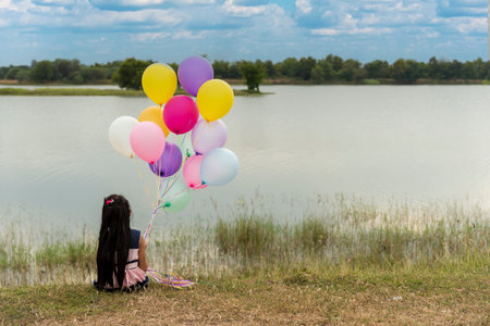 Cheerful beauty woman holding balloons relax sitting under big tree in green park with happiness. Woman Hands holding vibrant air balloons play on birthday party happy time summer on sunshine outdoorの写真素材