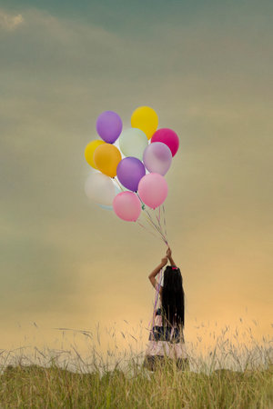 Cheerful beauty woman holding balloons relax sitting under big tree in green park with happiness. Woman Hands holding vibrant air balloons play on birthday party happy time summer on sunshine outdoorの写真素材