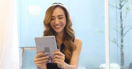 Happy asian woman video call web conference sitting on cozy sofa in living room at home. Female asian people use smart tablet headphones online call telecommunications from home watching videoの写真素材