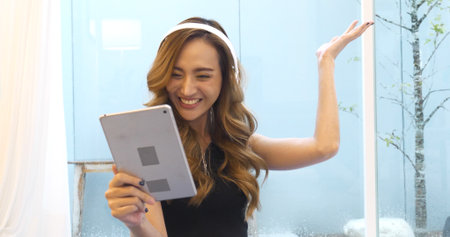 Happy asian woman video call web conference sitting on cozy sofa in living room at home. Female asian people use smart tablet headphones online call telecommunications from home watching videoの写真素材