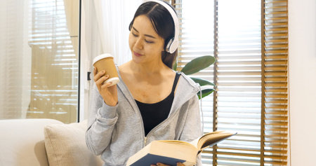 Happy asian woman use headphones listen to music reading book drink coffee cup sitting on cozy sofa in living room at home. Female asian people work at home read book magazine happy reader lifestyleの写真素材
