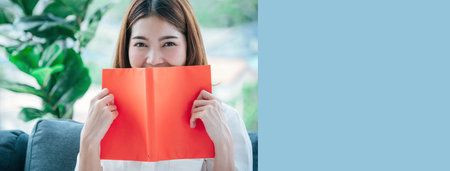 Banner Relax asian woman reading book sitting on sofa in living room holding book. Banner Young woman reading open book leisure mind. Happiness beauty person smiling face happy time with copy spaceの写真素材