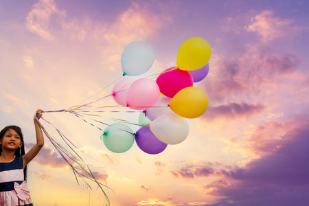 Cheerful cute girl holding balloons running on green meadow violet purple pastel sky happiness. Hands holding vibrant air balloons play on birthday party happy times summer on pueple sky outdoorの写真素材