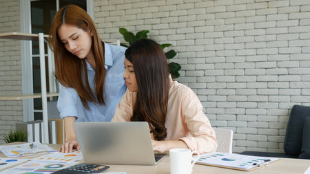 Two asian Female freelance reading financial graph charts Planning analyzing marketing data. Asian people team working office firm. Two businesswomen team meeting using laptop at company office desk.の写真素材