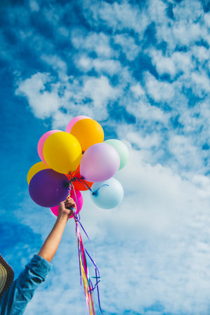 Vertical Cheerful cute girl holding balloons running on green meadow white cloud blue sky with happiness. Hands holding vibrant air balloons play on birthday party happy times sunlight outdoorの写真素材