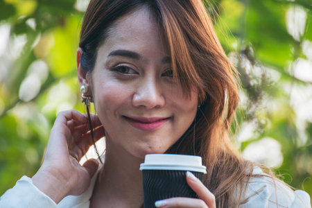 Woman drinking coffee hands holding hot disposable cup in green park. Happy Relax beautiful asian woman smiling face standing outdoors garden. Young women enjoy nature morning Freedom Lifestyle.の写真素材