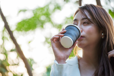Woman drinking coffee hands holding hot disposable cup in green park. Happy Relax beautiful asian woman smiling face standing outdoors garden. Young women enjoy nature morning Freedom Lifestyle.の写真素材