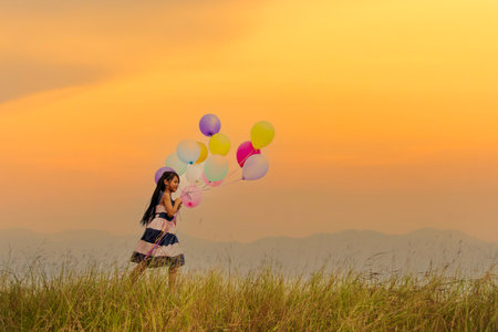 Cheerful cute girl holding balloons running on green meadow sunrise golden vibrant sky happiness....