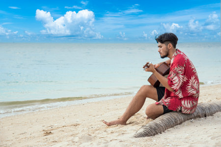 Young men playing acoustic guitar on the beach. Happy man person playing acoustic guitar music instrument seaside sitting on sand tropical beach island. Asian musician Happy guitarist hobby lifestyleの写真素材