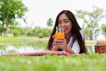 Asian Women picnic in green park smilng face hands holding glass of orange juice fresh fruit cool summer drinking. Beautiful women laydown picnic mat fruit basket hands holding healthy drinking glassの写真素材