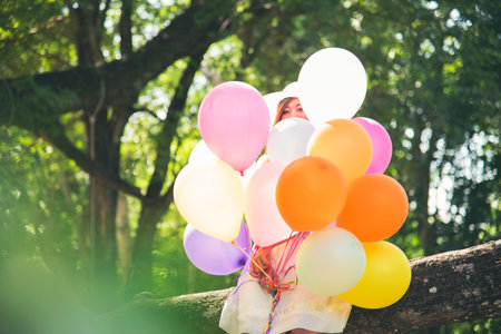 Cheerful beauty woman holding balloons relax sitting under big tree in green park with happiness. Woman Hands holding vibrant air balloons play on birthday party happy time summer on sunshine outdoorの写真素材